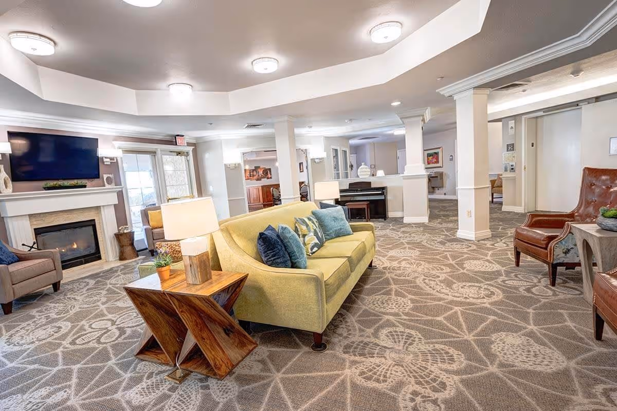 A spacious and well-lit living room area in an assisted living facility featuring a yellow sofa with blue and patterned cushions, a wooden side table with a lamp and small plant, a fireplace with a mounted flat-screen TV above it, and additional seating including a brown leather chair and a gray armchair. The room has patterned carpet flooring, white walls, ceiling lights, and a piano in the background.