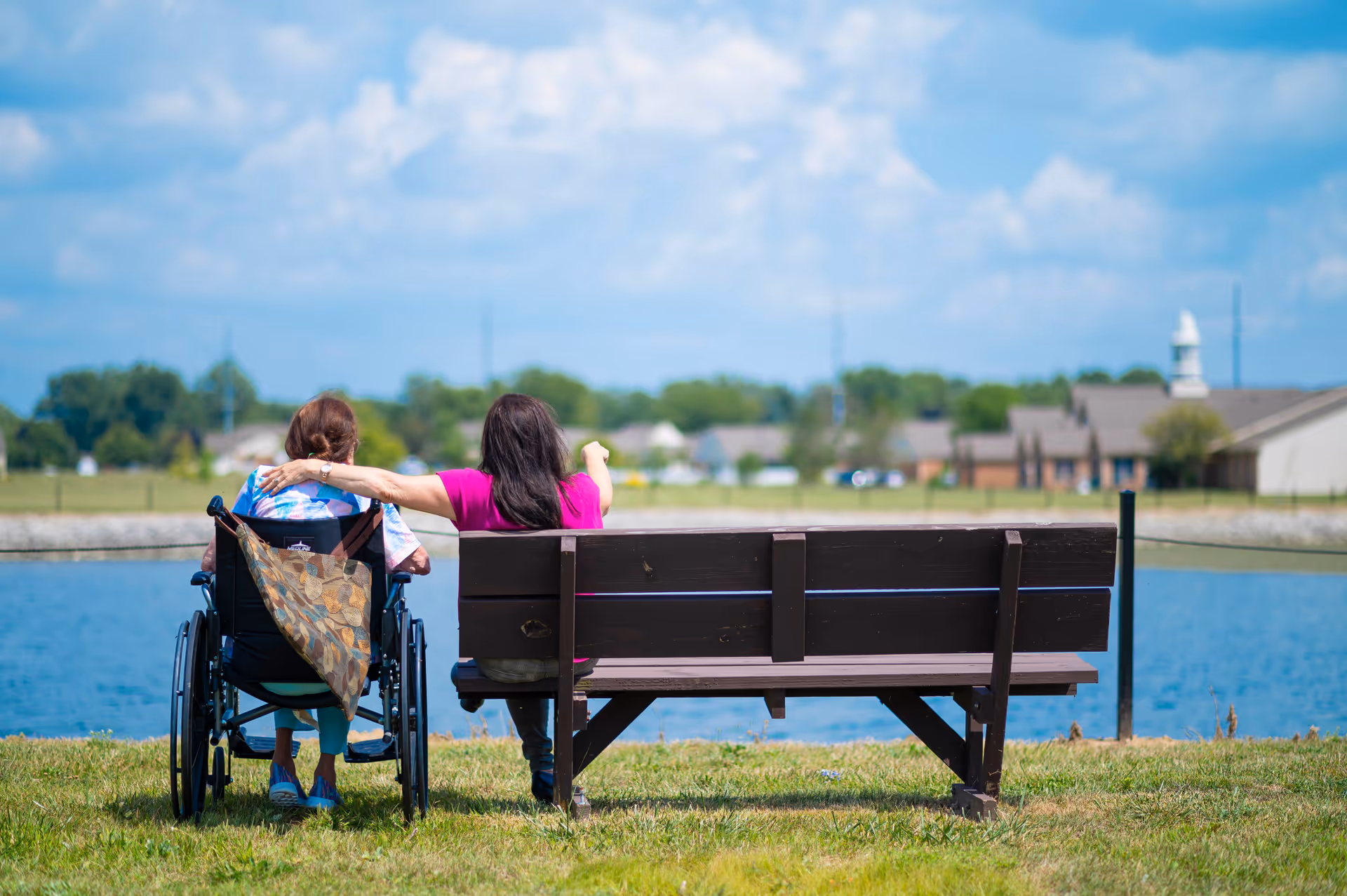 Two women sitting outdoors by a pond, one in a wheelchair and the other on a wooden bench. The woman on the bench has her arm around the woman in the wheelchair and is pointing towards something in the distance. The background shows a grassy area, houses, and a partly cloudy sky.