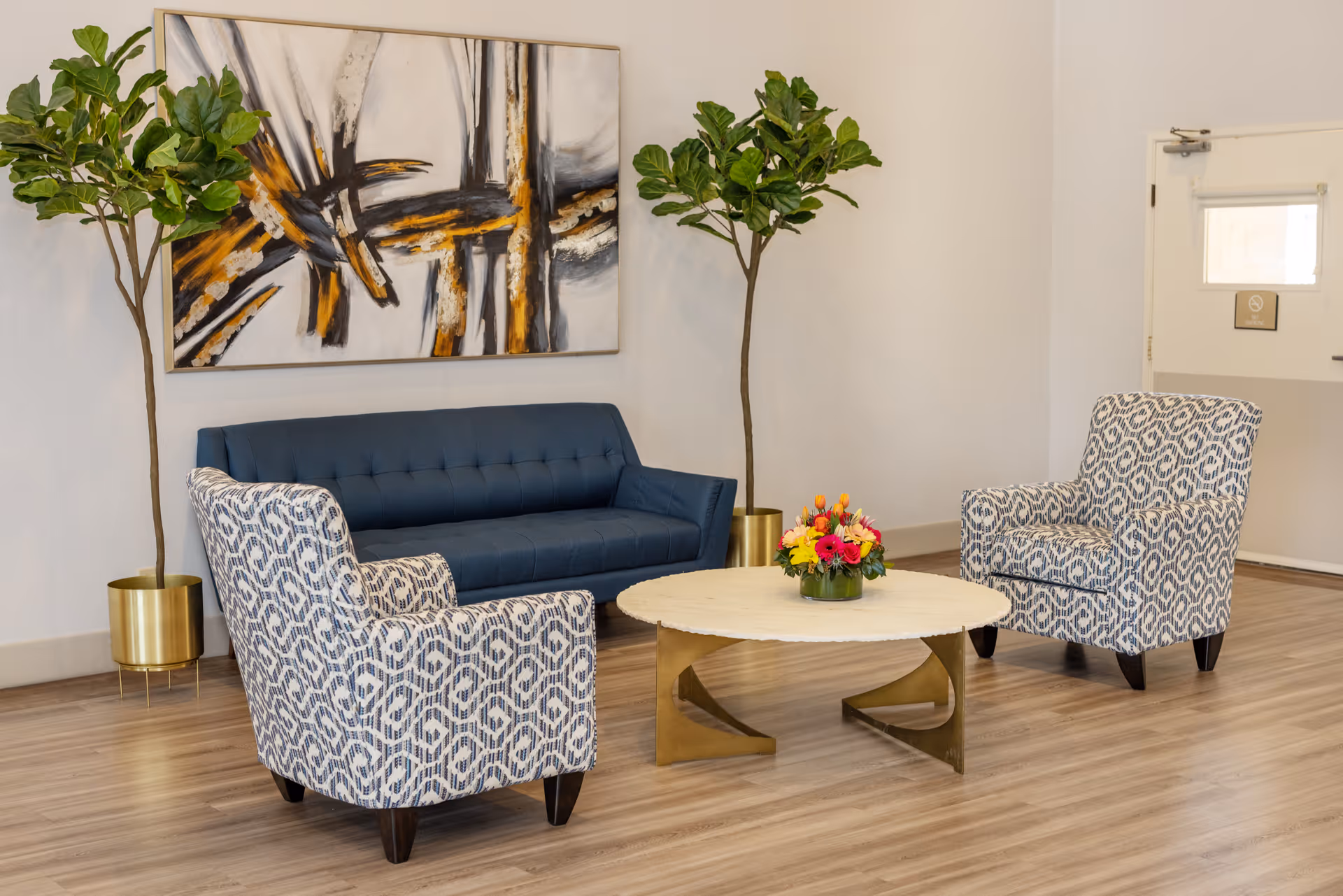 A modern seating area with a blue sofa, two patterned armchairs, a round coffee table with a flower arrangement, two tall potted plants, and an abstract painting on the wall.