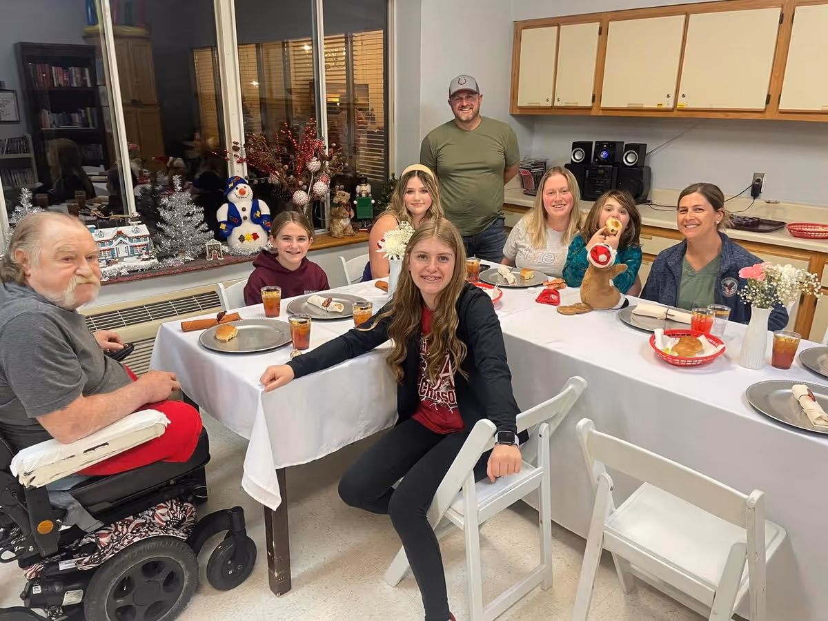 A group of residents and visitors seated around a decorated table in a communal dining area, smiling at the camera.