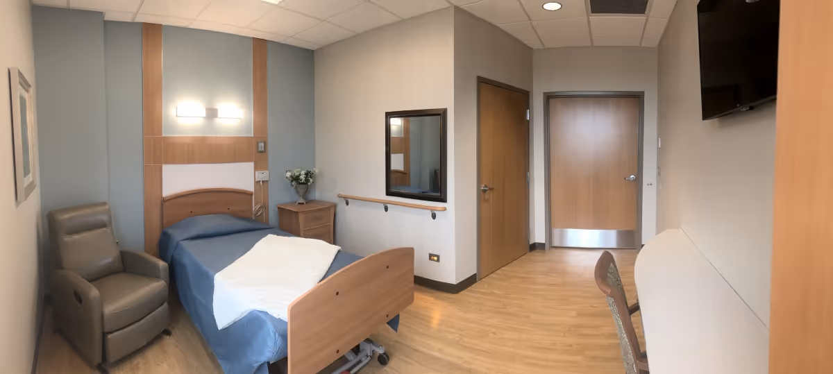 A single bedroom in a senior living facility featuring a hospital-style bed with blue bedding and a white blanket, a brown recliner chair, a wooden nightstand with a vase of flowers, a wall-mounted light fixture above the bed, a large mirror on the wall, two closed wooden doors, a wall-mounted flat screen TV, and a chair at a long desk or counter along the opposite wall. The room has light-colored walls and wood flooring.