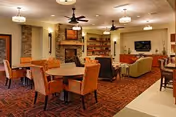 A warm and inviting living room area in a senior living facility featuring multiple seating arrangements including orange upholstered chairs around wooden tables, beige sofas, and armchairs. The room has a stone fireplace with a mounted TV above it, ceiling fans, and shelves with decorative items. The carpet has a red and brown pattern, and the lighting is soft with hanging fixtures.