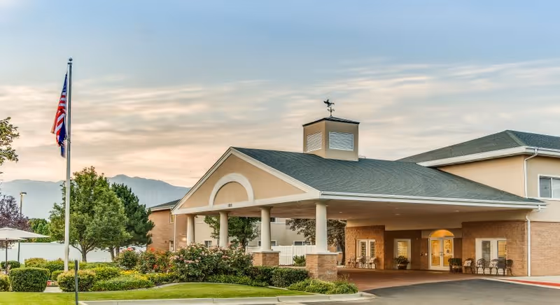 Covered entrance of a senior living building with a porte-cochère, landscaped grounds, and an American flag.