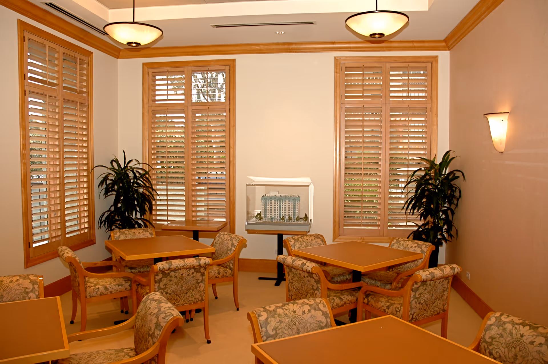A small dining or common area with several square tables and upholstered chairs with floral patterns. The room has three large windows with wooden shutters, two potted plants in the corners, and a model of a building displayed on a small table against the wall. The walls are light-colored with wooden trim, and there are ceiling and wall-mounted light fixtures providing warm lighting.
