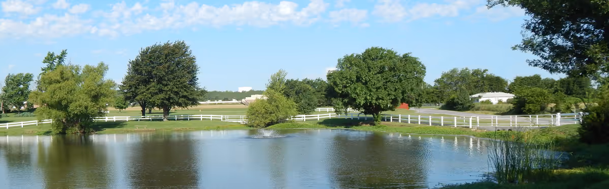A peaceful outdoor scene at Franciscan Villa featuring a small pond with a water fountain in the center, surrounded by green grass, trees, and white fencing under a partly cloudy blue sky.