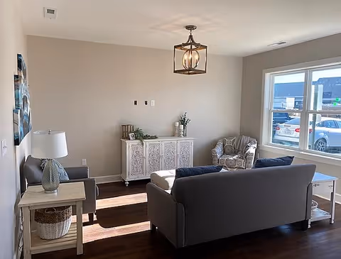 A bright living room with a large window showing parked cars outside. The room features a gray sofa, a patterned armchair, a white side table with a lamp, a decorative white cabinet against the wall, and a modern ceiling light fixture. The walls are painted beige and the floor is dark wood.