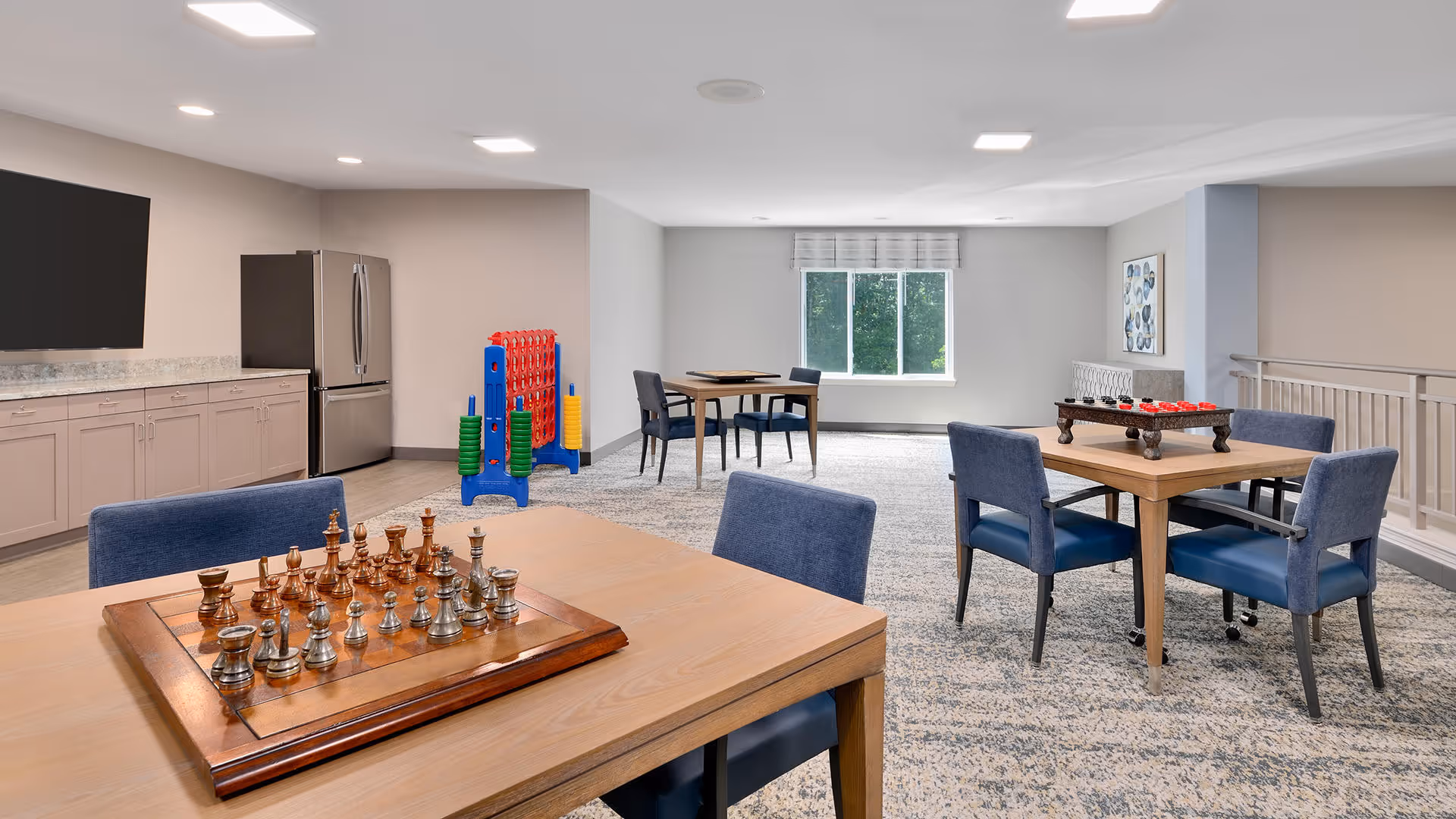 A bright and spacious common area with multiple wooden tables and blue upholstered chairs. One table has a chess set, another has a checkers game. In the background, there is a large window letting in natural light, a stainless steel refrigerator, cabinets, and a large Connect Four game. The room has a light-colored carpet and neutral walls.