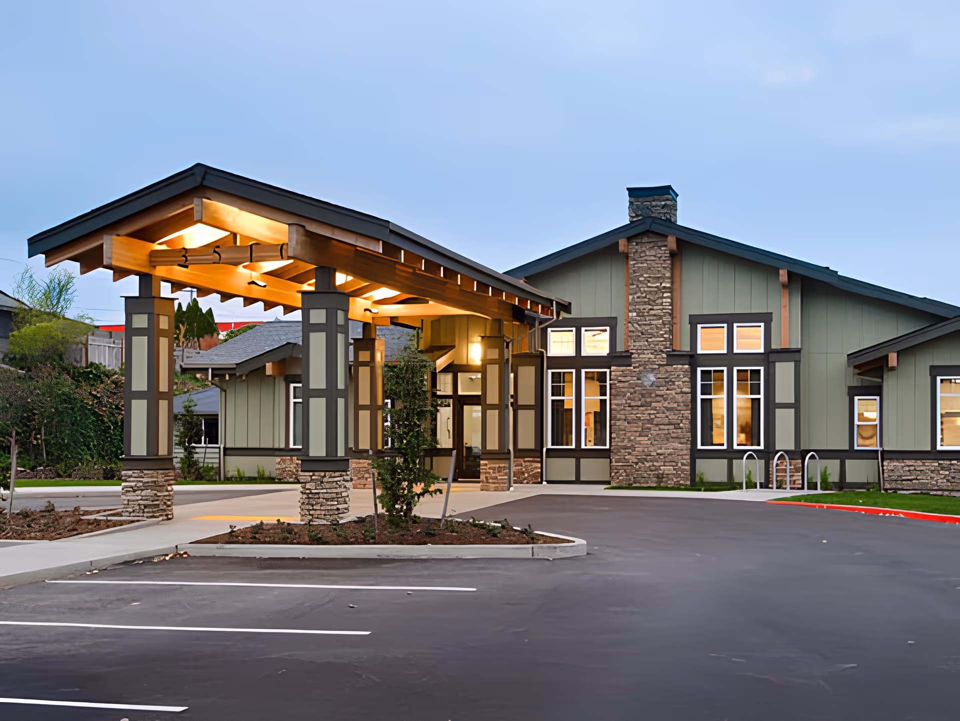 Exterior view of The Arbor at Bremerton facility during dusk, showing a covered entrance with warm lighting, stone and wood architectural details, large windows, and a paved parking area in front.