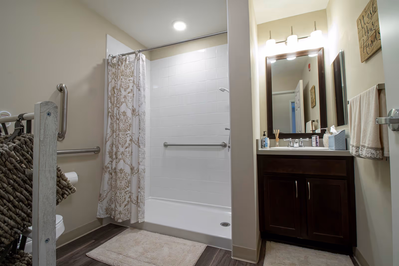 A bathroom with a walk-in shower featuring white tiled walls and a decorative shower curtain. There are grab bars installed inside the shower and on the adjacent wall. A dark wood vanity with a white countertop and a large mirror above it is visible, along with a towel rack holding a beige towel. The floor is covered with a light-colored bath mat and dark wood-style flooring.