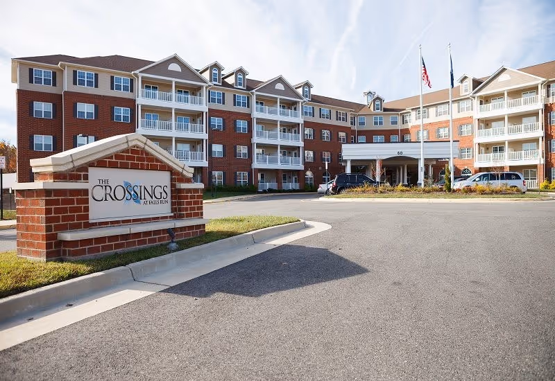 Exterior front view of The Crossings At Falls Run senior living facility, showing a multi-story brick building with balconies, a driveway, parked cars, and a sign with the facility name in the foreground.