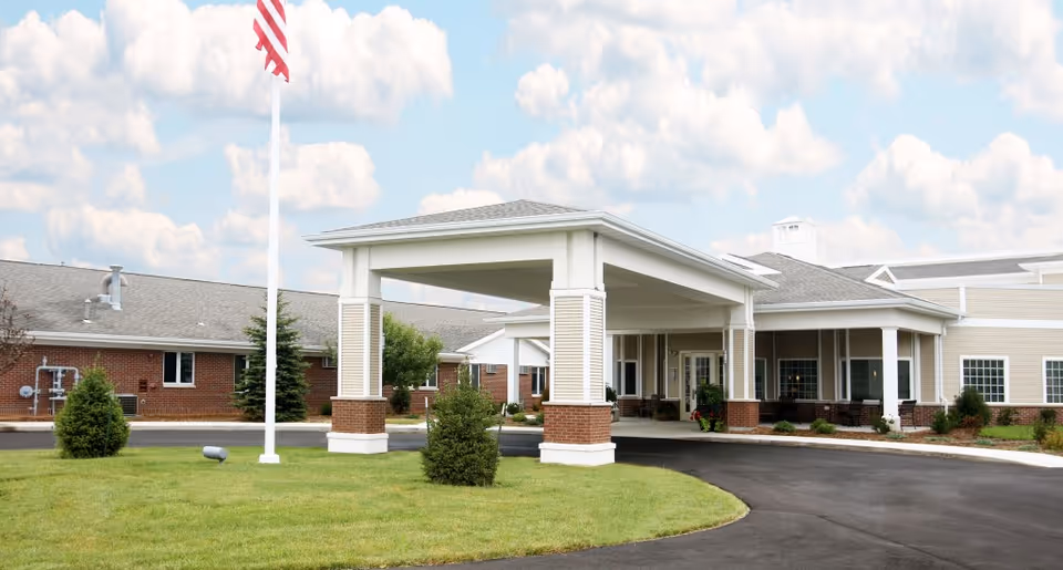 Entrance canopy and driveway of a single-story senior care facility with an American flag and landscaped lawn.