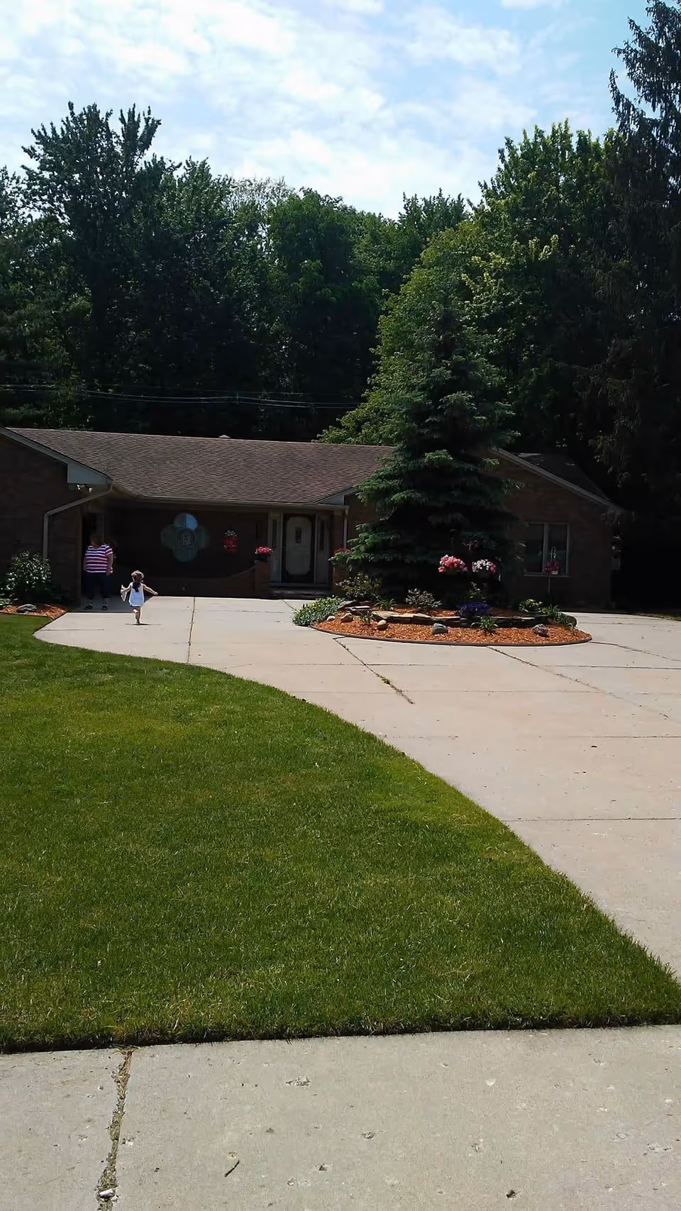 A single-story brick building with a sloped roof, surrounded by trees and greenery. There is a concrete driveway leading up to the entrance, which has a small garden bed with a large evergreen tree and flowers in front. Two people, an adult and a child, are walking on the driveway near the entrance.