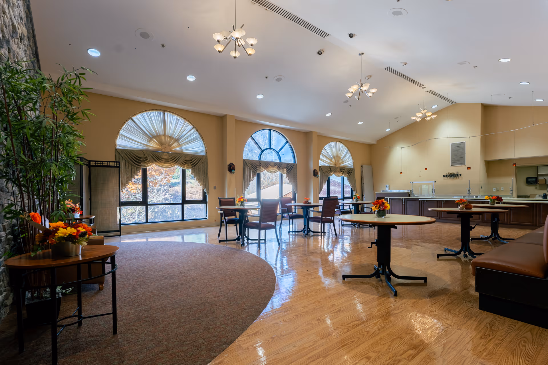 A spacious and well-lit dining area in a senior living facility with several round tables and chairs arranged neatly. Large arched windows with decorative curtains allow natural light to fill the room. The floor is a combination of polished wood and carpet, and there are ceiling lights and hanging fixtures providing additional illumination. A counter area is visible in the background, likely for food service, and there are small flower arrangements on the tables and side tables.