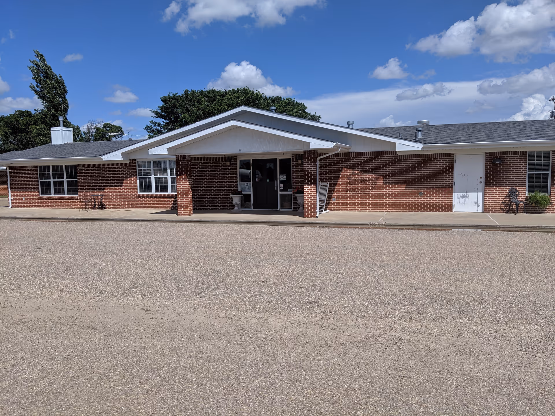 Front exterior of a single-story red brick nursing home building with a covered main entrance and paved driveway under a blue sky.