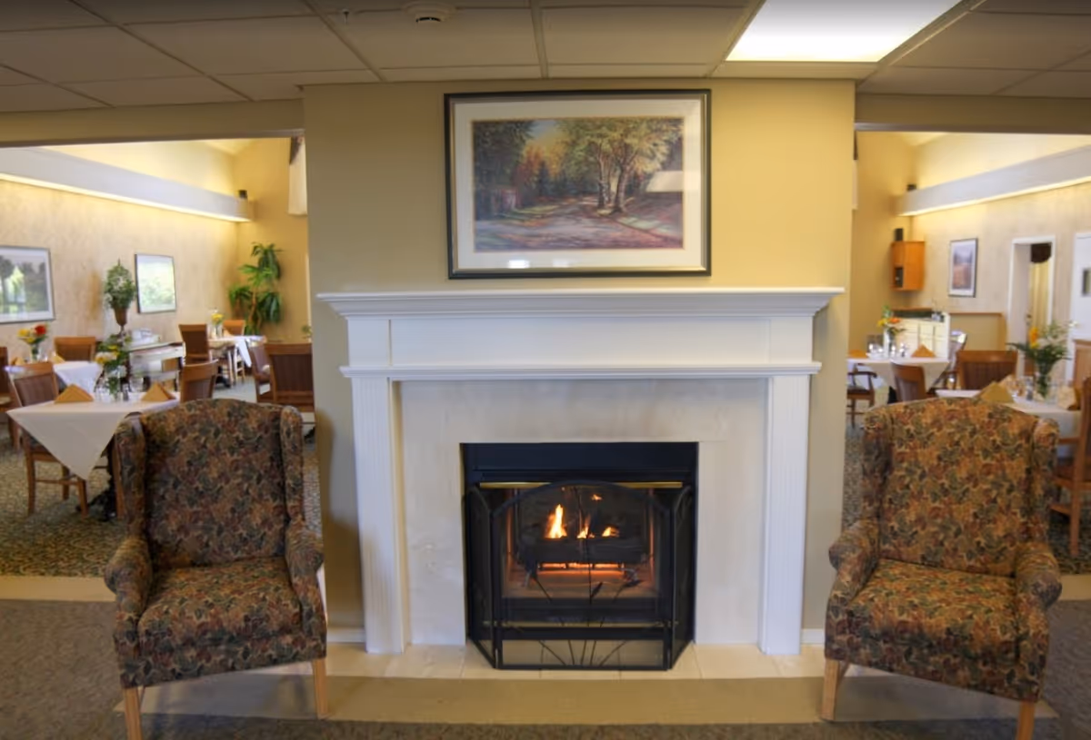 A cozy common area with a lit fireplace flanked by two patterned armchairs and dining tables visible in the background.