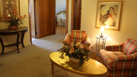 A cozy sitting area with two plaid armchairs and a wooden coffee table adorned with a floral arrangement. A small side table with a lamp and a framed painting on the wall add to the warm ambiance. In the background, an open doorway reveals a bedroom with a floral bedspread.