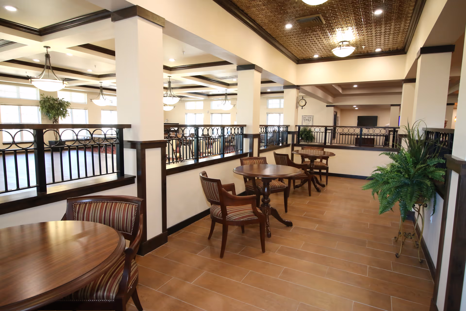 Interior view of a senior living facility lounge area with round wooden tables and striped cushioned chairs along a tiled floor. The space features white walls with dark wood trim, decorative ceiling tiles, hanging light fixtures, and potted plants. A railing separates this seating area from a larger common area with windows letting in natural light.