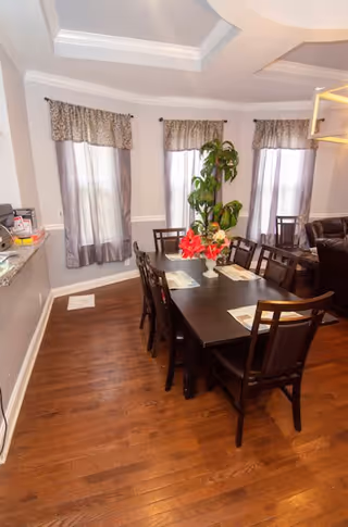 A dining area featuring a large table surrounded by chairs, with floral decorations and large windows.