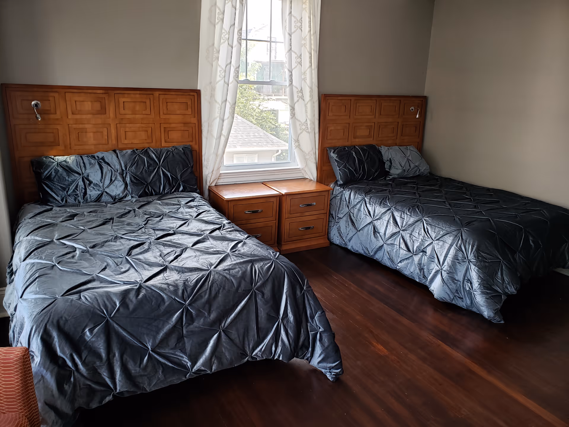 A bedroom with two beds featuring wooden headboards and dark gray bedding. Between the beds is a wooden nightstand with two drawers. A window with white patterned curtains is centered on the wall behind the nightstand, allowing natural light into the room. The floor is dark wood.