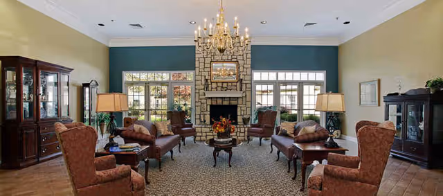A spacious and elegant living room with a stone fireplace centered on a teal accent wall. The room features multiple upholstered armchairs and sofas arranged around a central wooden coffee table with a floral arrangement. Large windows on either side of the fireplace allow natural light to fill the space. Two wooden display cabinets and two table lamps with beige shades are positioned on either side of the room. A chandelier hangs from the ceiling, illuminating the area.