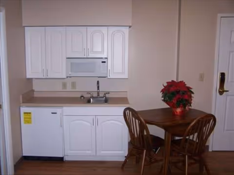 Compact kitchenette with white cabinets, microwave, sink, mini-fridge, and a wooden dining table with a poinsettia.