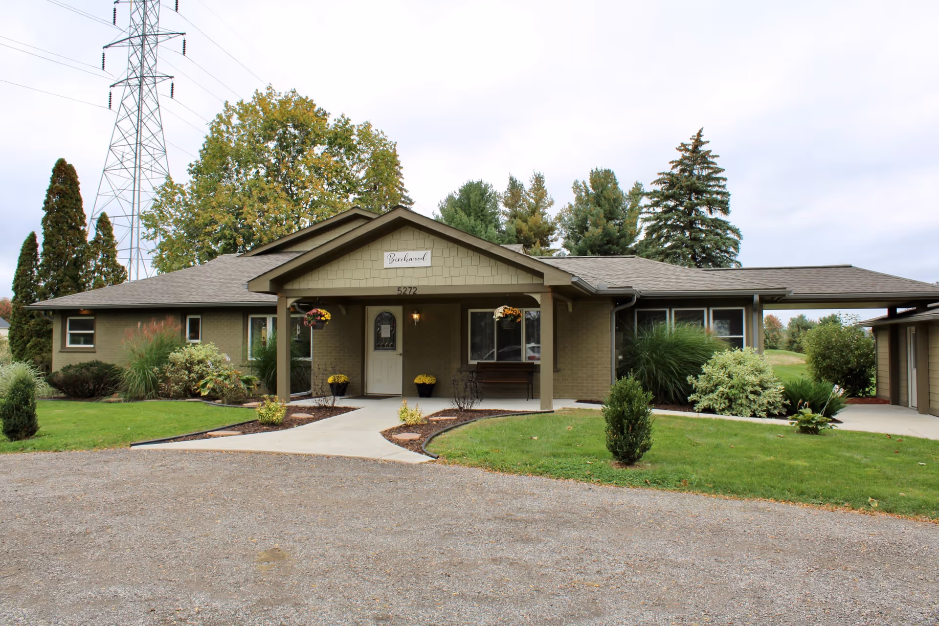 Front exterior view of Birchwood Gardens facility showing a single-story building with a covered entrance, hanging flower pots, a bench, and well-maintained landscaping with green grass and shrubs.