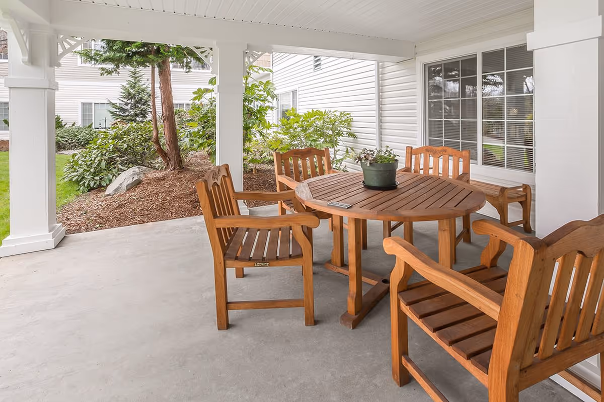 Covered outdoor patio area with a round wooden table and five wooden chairs. A potted plant is placed on the table. The patio overlooks a garden with shrubs, trees, and mulch, adjacent to a white building with windows.