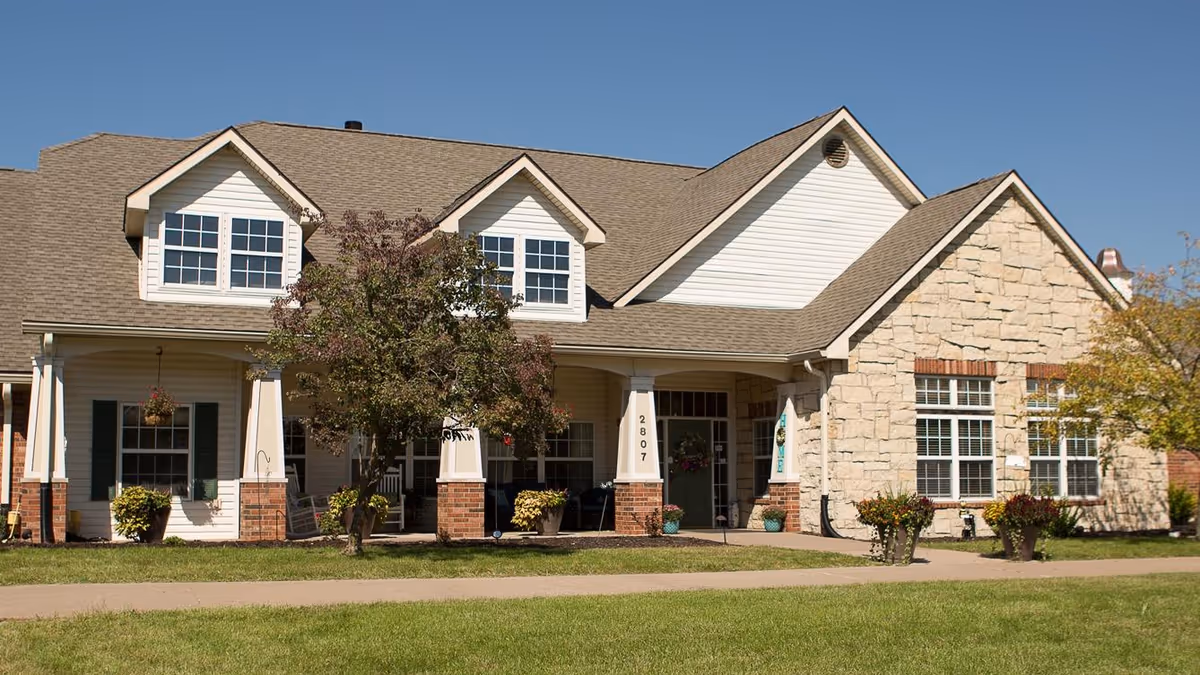 Exterior view of a single-story residential building with a combination of white siding and stone facade, featuring multiple windows, a covered porch with columns, potted plants, and a tree in front under a clear blue sky.