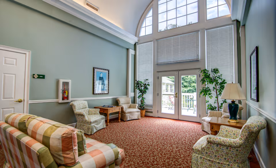 A bright and cozy sitting area with high arched windows and glass doors leading outside. The room features a patterned red carpet, pastel-colored armchairs and sofas, wooden side tables, potted plants, a table lamp, and framed artwork on the light green walls.