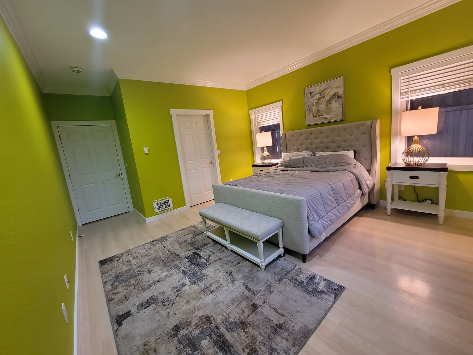 A bedroom with bright green walls, light wood flooring, and a large bed with a tufted headboard. The bed is covered with a gray comforter and has two pillows. On either side of the bed are white nightstands with table lamps. There are two windows with blinds behind the nightstands. A patterned area rug is placed in front of the bed, and a small bench is positioned at the foot of the bed. Two white doors are visible on the left wall.