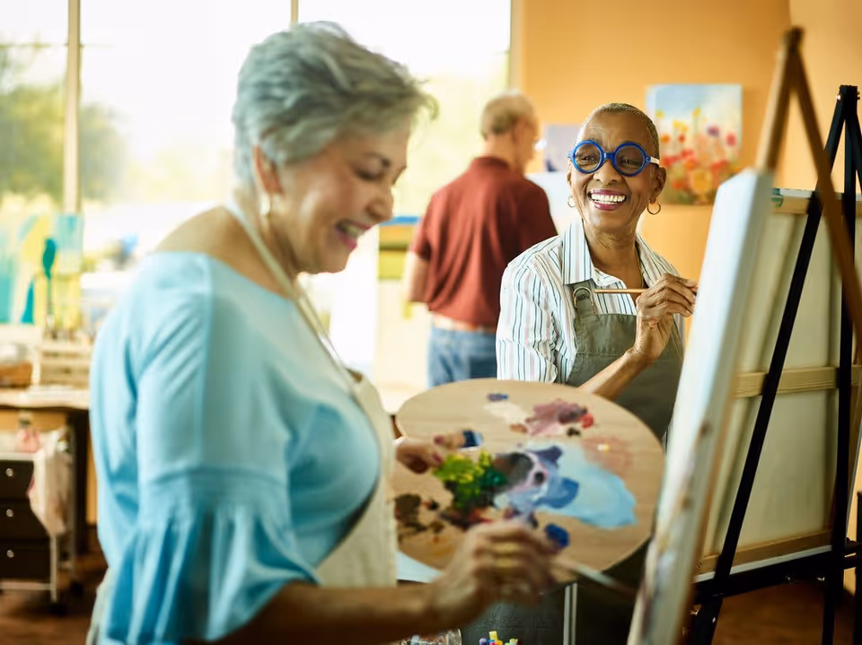 Two elderly women smiling and painting on canvases in an art studio, with a man in the background also engaged in painting. One woman is holding a paint palette and brush, while the other is wearing blue round glasses and an apron.