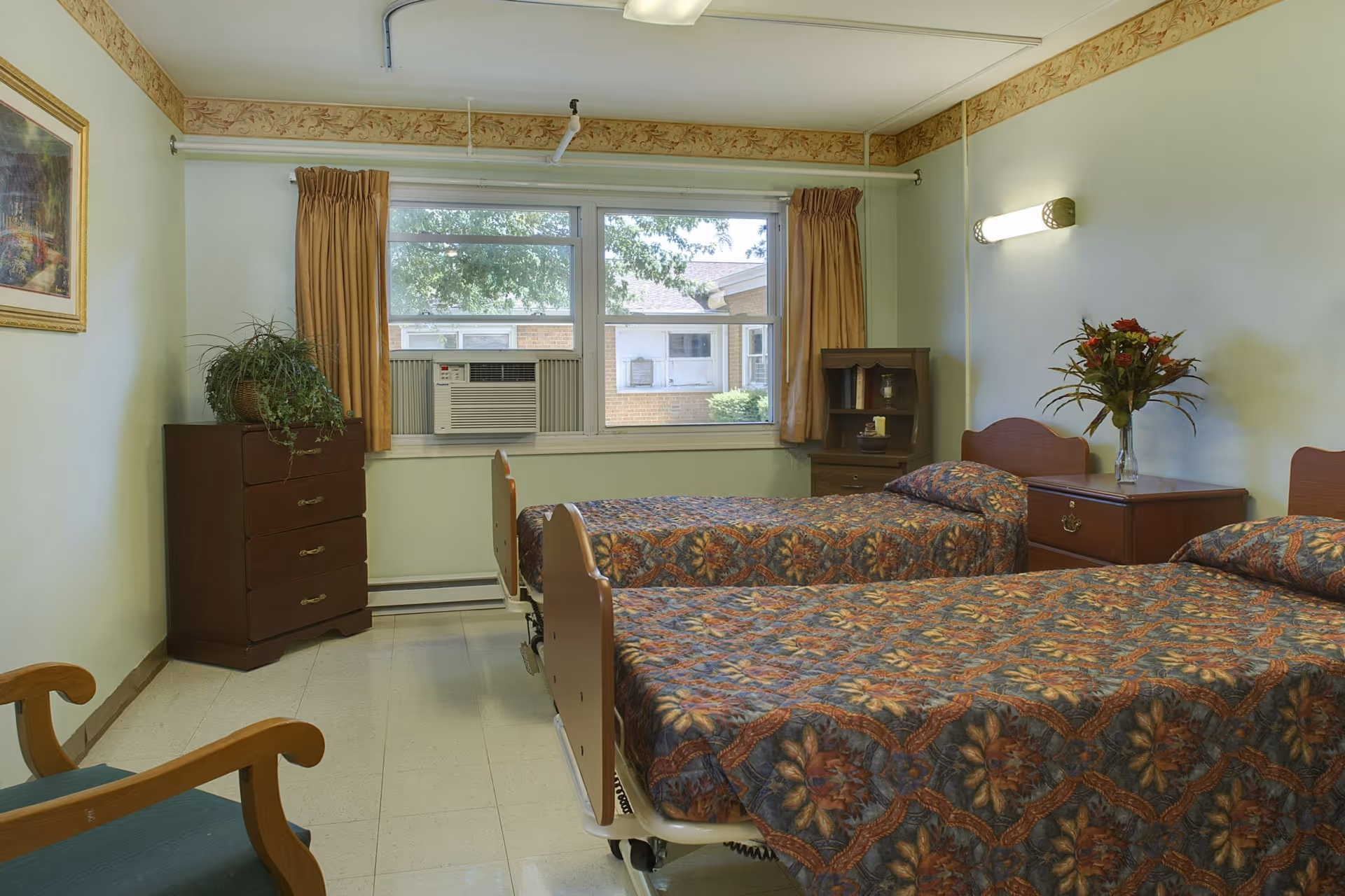 Bright bedroom with two twin beds, patterned bedspreads, a dresser, window air conditioner and a vase of flowers on a nightstand.