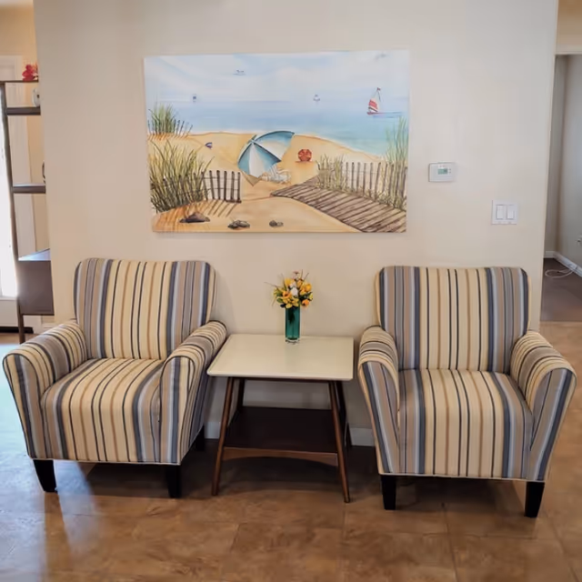 Two striped armchairs flank a small table with a vase of flowers beneath a beach-themed painting on a light-colored wall.