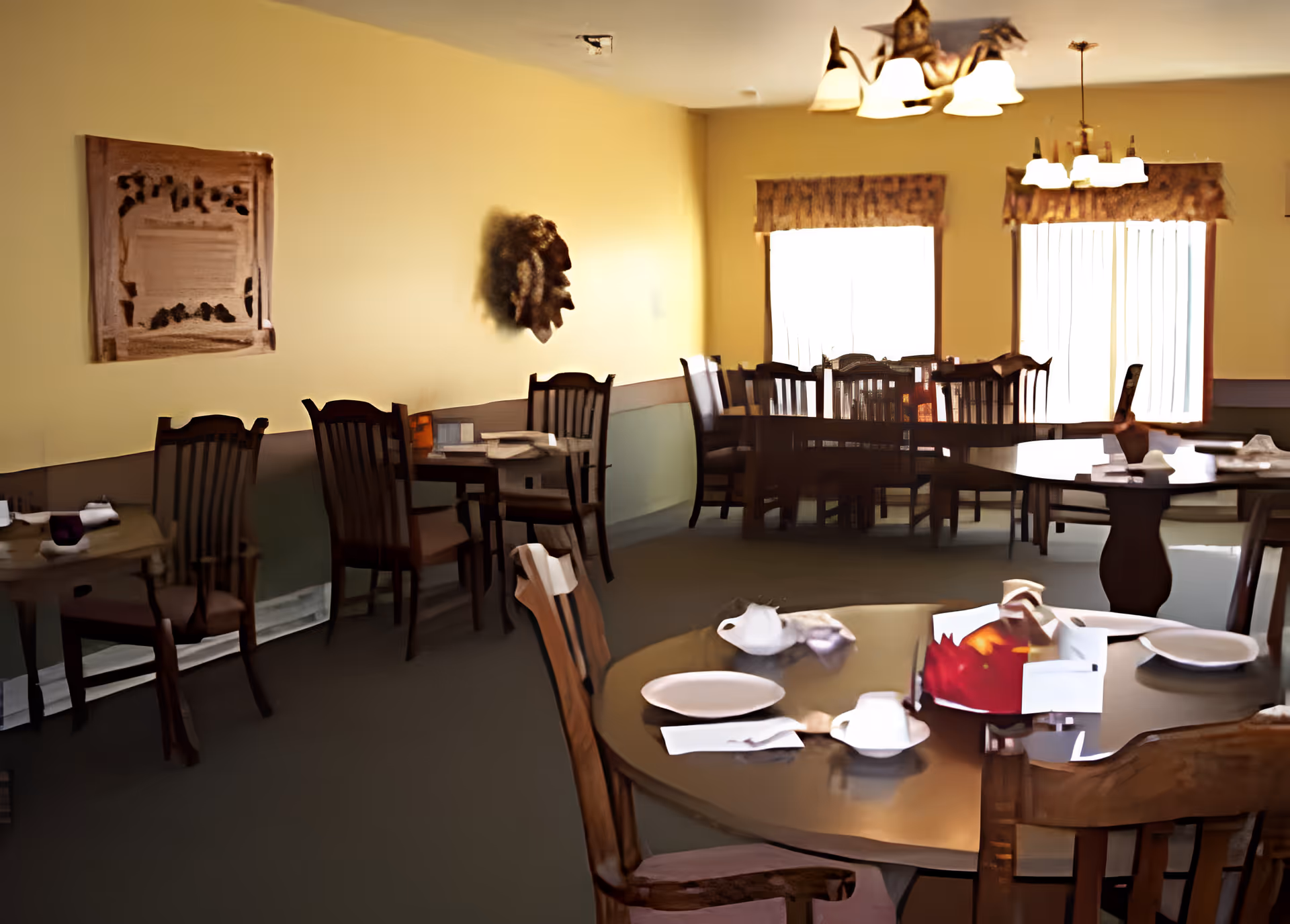 Interior view of a dining room with multiple wooden tables and chairs. The tables are set with plates, cups, and napkins. The room has yellow walls, two windows with curtains, and ceiling light fixtures.