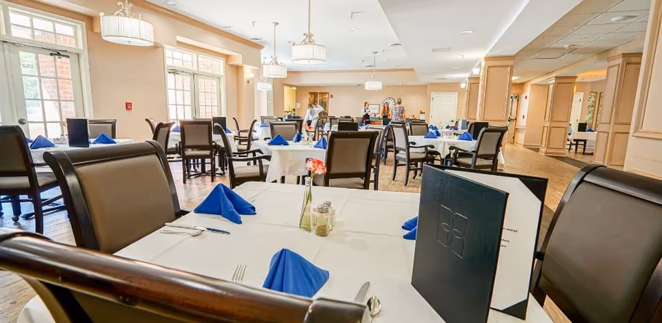 A bright dining room with multiple tables covered in white tablecloths, each set with blue folded napkins, silverware, and a small flower vase. Several chairs surround the tables, and large windows allow natural light to fill the room. In the background, a few people are standing near the far end of the room.