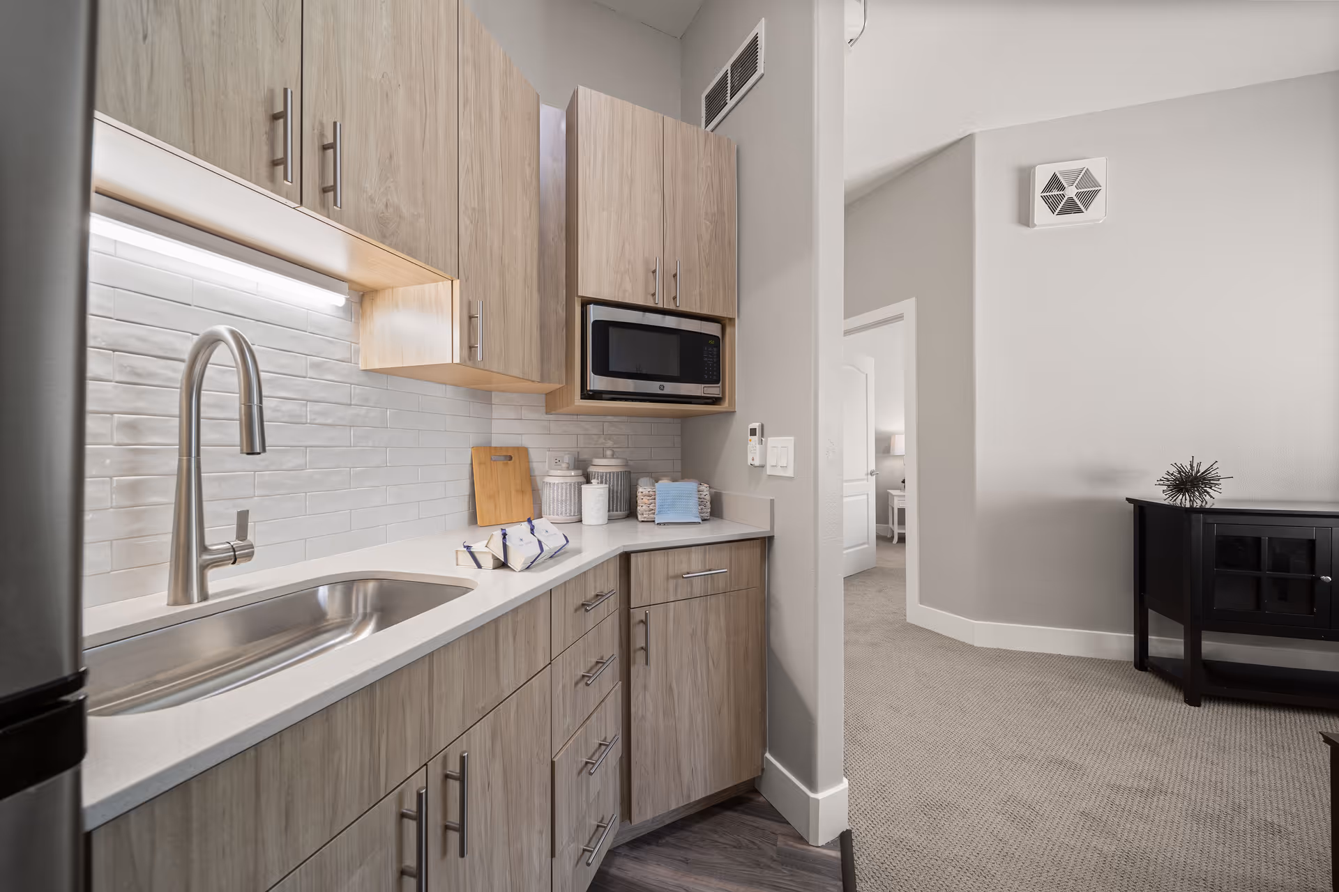 Modern kitchen area with light wood cabinets, a stainless steel sink and faucet, a built-in microwave, and white subway tile backsplash. The kitchen counter has decorative jars, a cutting board, and a few wrapped items. Adjacent to the kitchen is a carpeted hallway leading to a bedroom with a white door and a bedside table with a lamp. A black console table with a decorative object is visible in the living area.