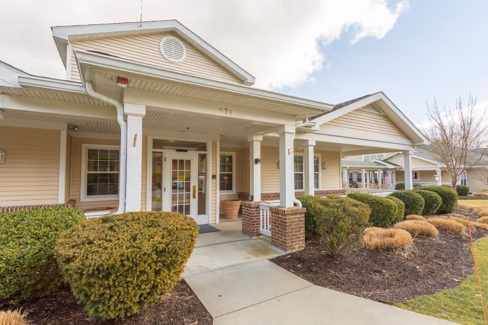 Entrance of a beige single-story senior living facility with a covered porch, white columns, glass doors, and landscaped shrubs.