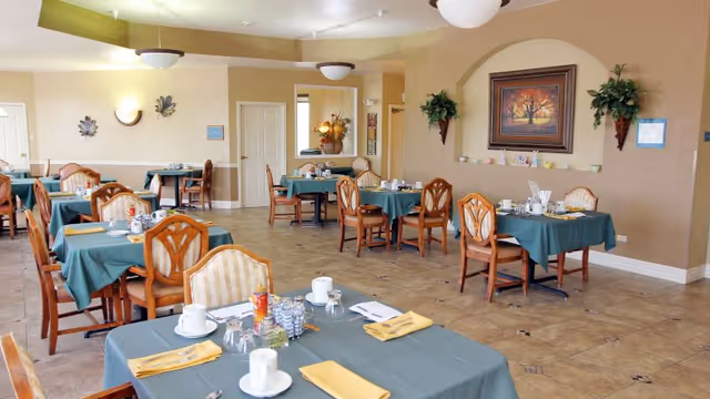A dining room in a senior living community with multiple tables covered in green tablecloths, each set with white cups, glasses, silverware, and yellow napkins. The room has beige walls, tile flooring, and decorative wall art including a framed painting and wall-mounted plants.