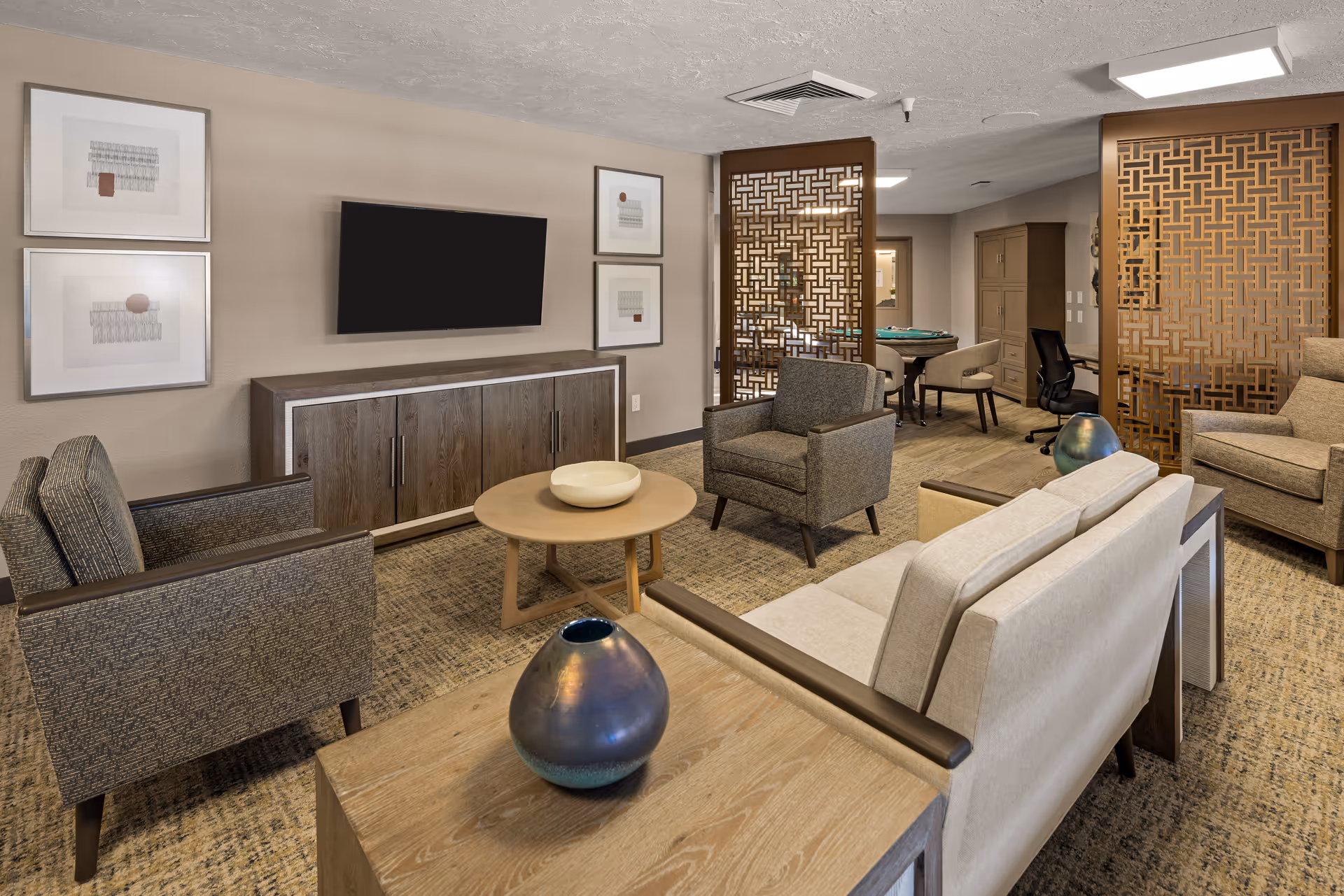 A cozy senior living common area with a beige sofa, two gray armchairs, a wooden coffee table with a decorative bowl, and a wooden side table with a blue vase. A flat-screen TV is mounted on the wall above a wooden cabinet. The room features decorative wooden lattice partitions and a carpeted floor.