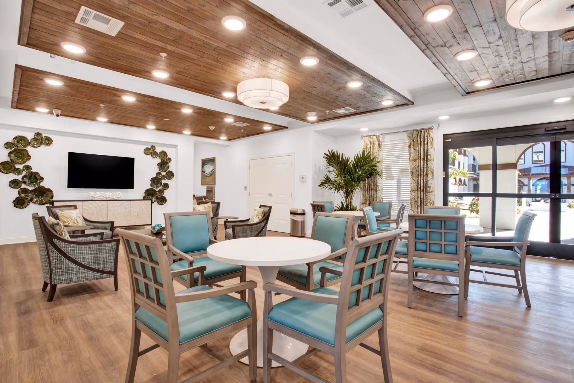 Bright communal lounge with round tables, teal-upholstered chairs, lounge seating, a wall-mounted TV, and large glass doors.