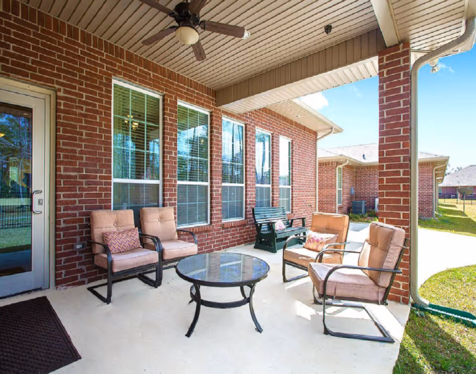 Covered outdoor patio area with brick walls, four cushioned chairs around a glass-top round table, a green bench with a cushion, ceiling fan, and a view of the grassy yard and neighboring buildings under a clear blue sky.