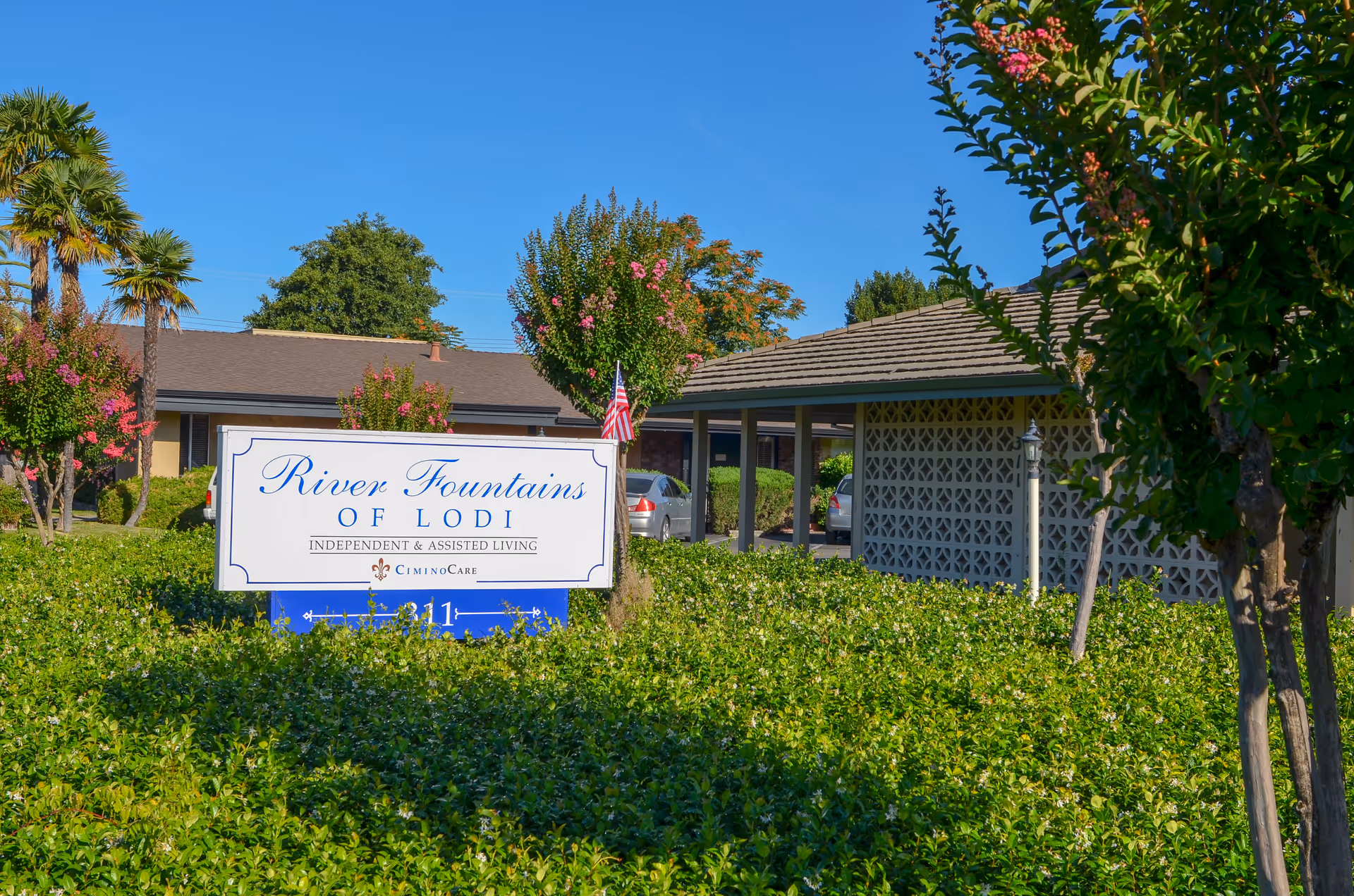Outdoor view of the entrance area of River Fountains of Lodi, an independent and assisted living facility, with a sign surrounded by green bushes and trees under a clear blue sky.