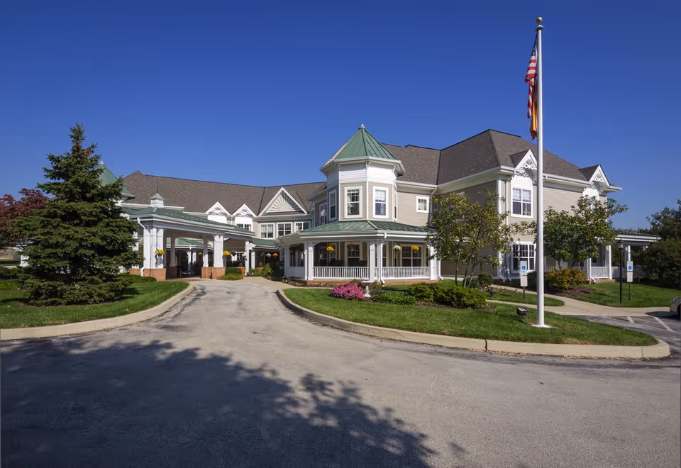 Exterior view of a large, elegant senior living facility building with a green roof and beige siding under a clear blue sky. The building features a covered entrance, a circular driveway, manicured lawns, trees, and an American flag on a flagpole in front.