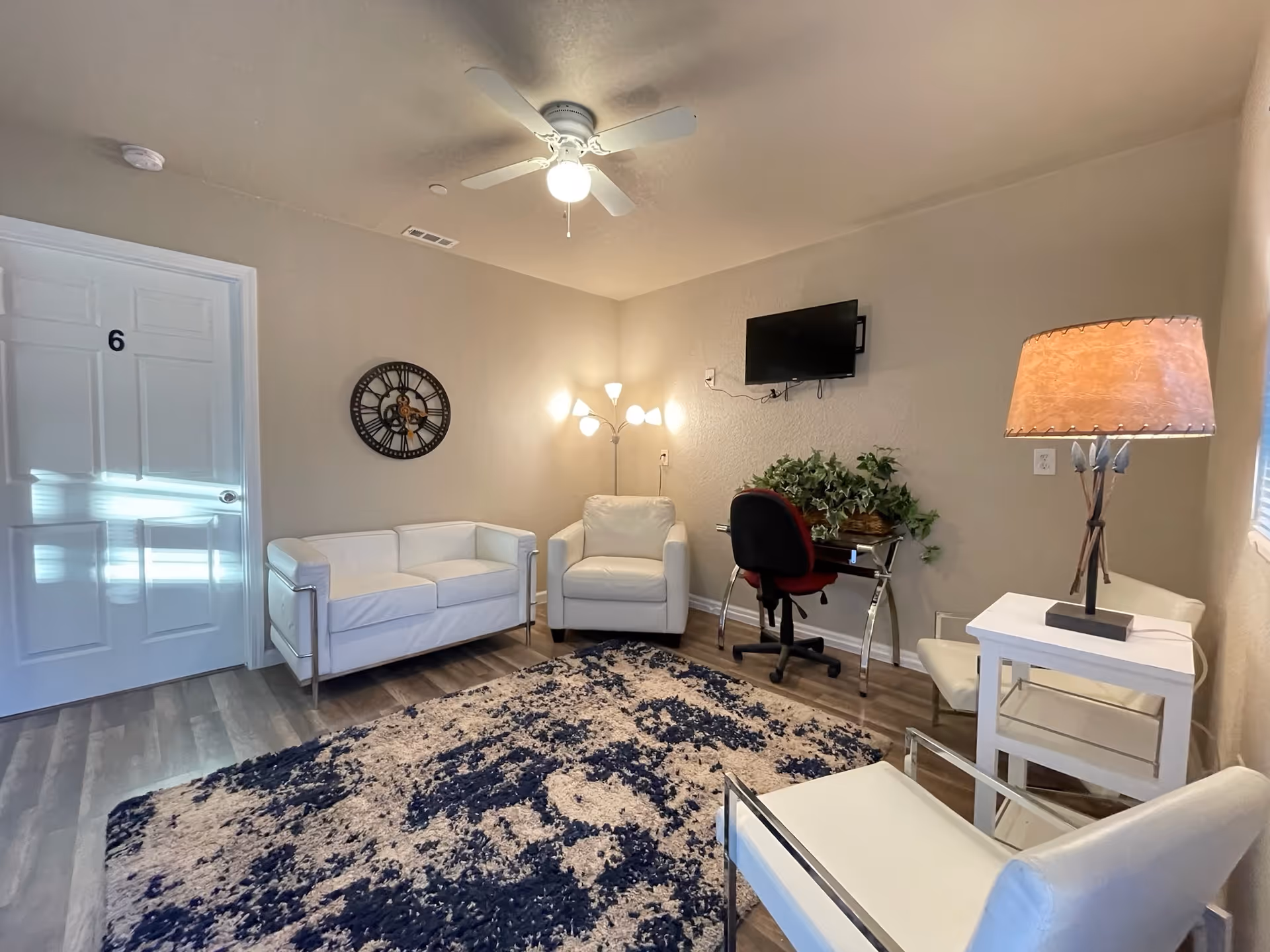 A cozy living room area with white leather chairs and a loveseat, a patterned rug, a wall-mounted TV, a desk with a red office chair and a plant, a floor lamp, and a table lamp on a small white side table. The room has beige walls, a ceiling fan with a light, and a door labeled with the number 6.