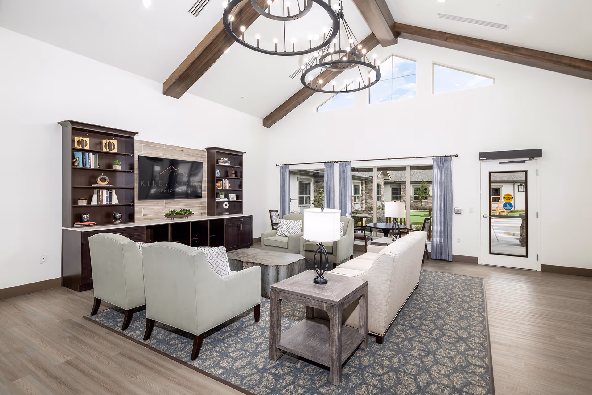 A bright and spacious senior living facility common area with high vaulted ceilings and exposed wooden beams. The room features a seating arrangement with two armchairs, a sofa, and a wooden coffee table on a patterned rug. There are two large circular chandeliers hanging from the ceiling. A large flat-screen TV is mounted on the wall between two dark wooden bookshelves filled with decorative items and books. Large windows with blue curtains provide a view of the outdoor courtyard. A door with a window and accessibility stickers is visible on the right side.
