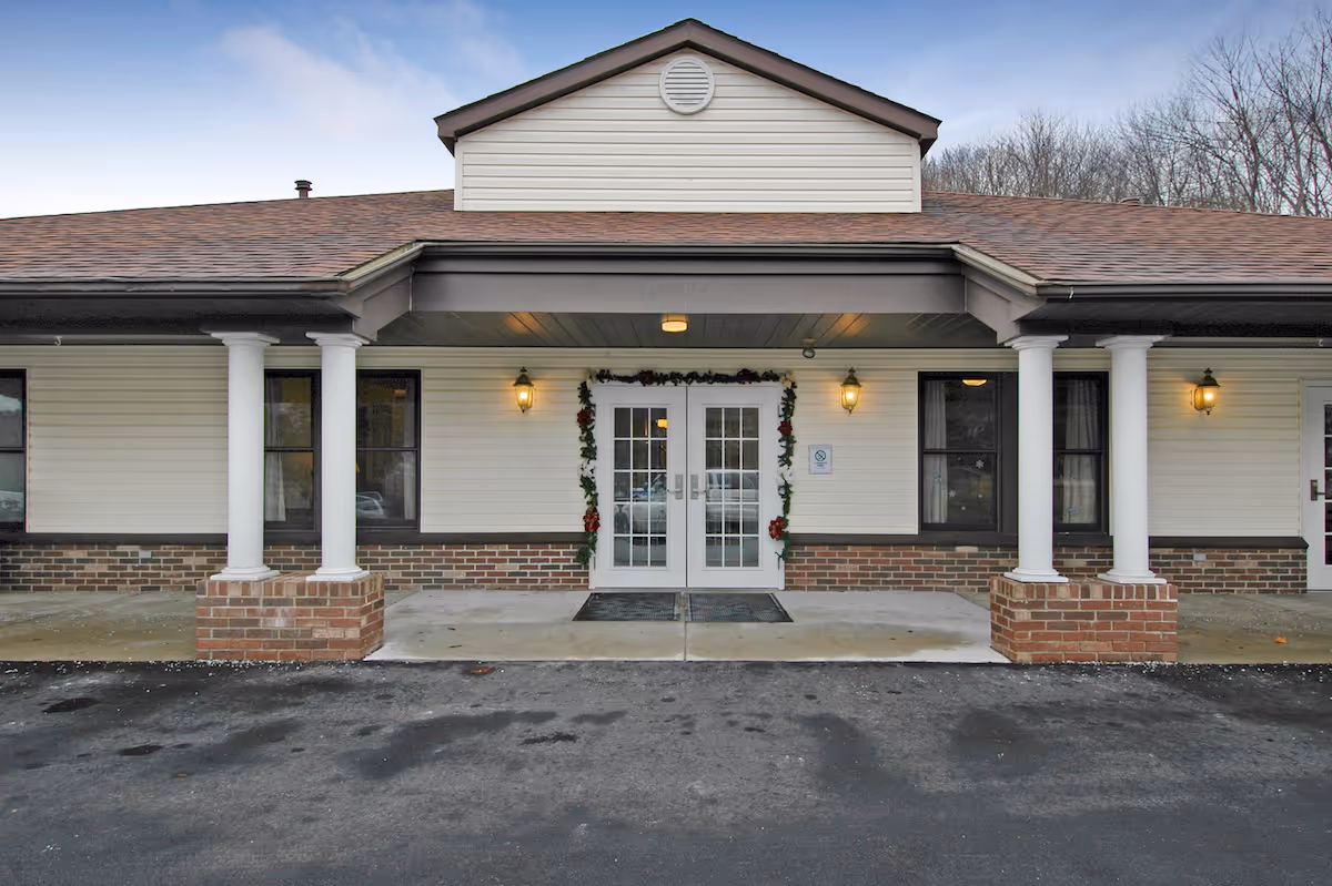 Front entrance of a single-story building with double glass doors flanked by white columns and brick bases, decorated with garland and exterior lights.