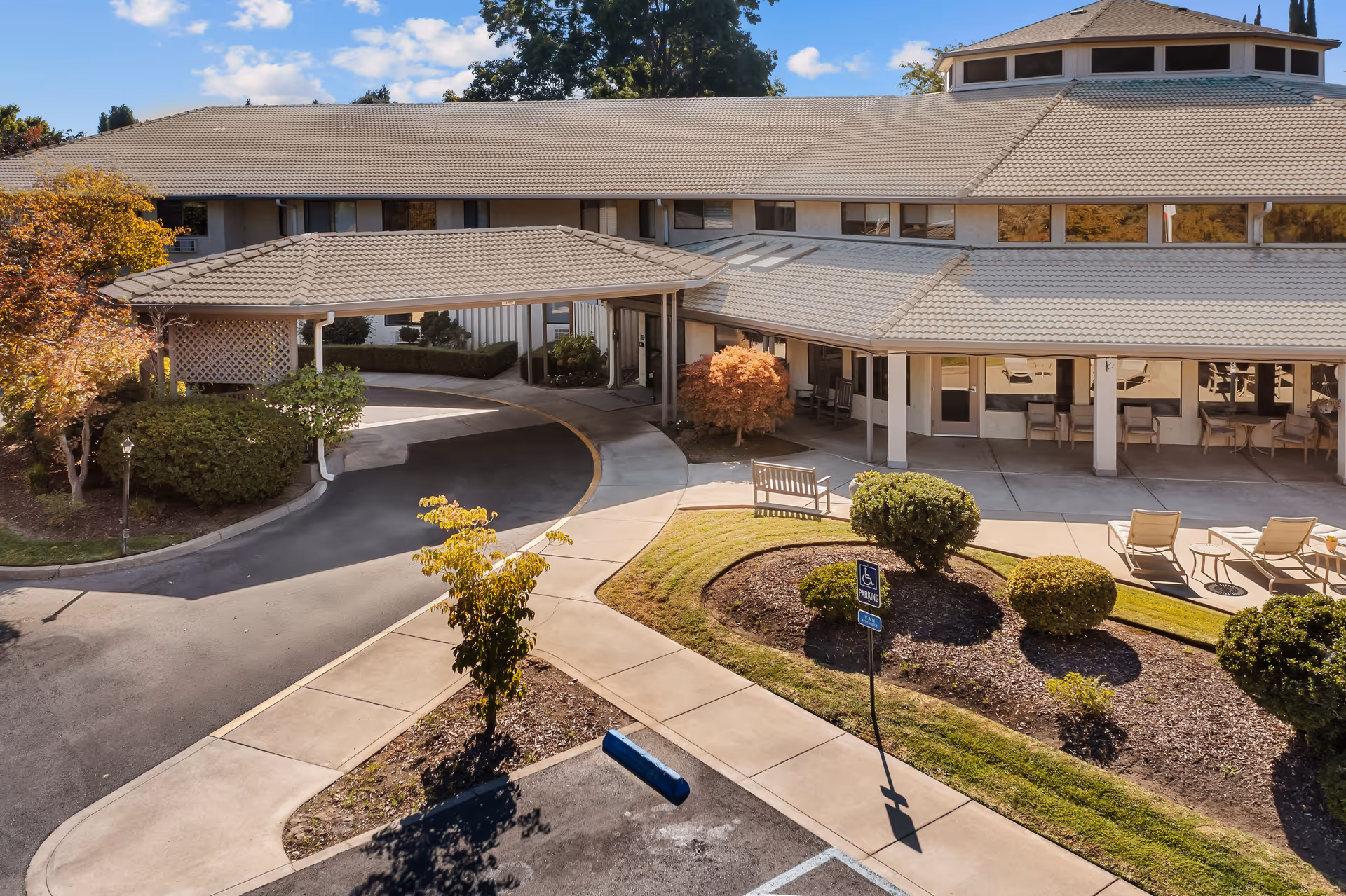 Exterior view of a senior living facility with a covered entrance, landscaped garden with bushes and small trees, a wheelchair accessible parking space, and outdoor seating including benches and lounge chairs under a covered patio.