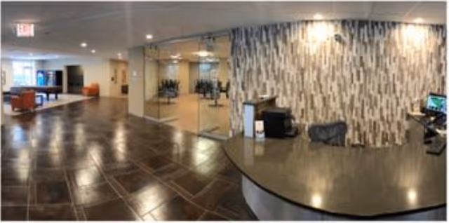 Reception area of a senior living facility with a curved front desk on the right, a patterned wall behind it, and a glass-walled room with chairs and plants visible. The floor is tiled with large dark tiles, and there are seating areas with orange chairs in the background.