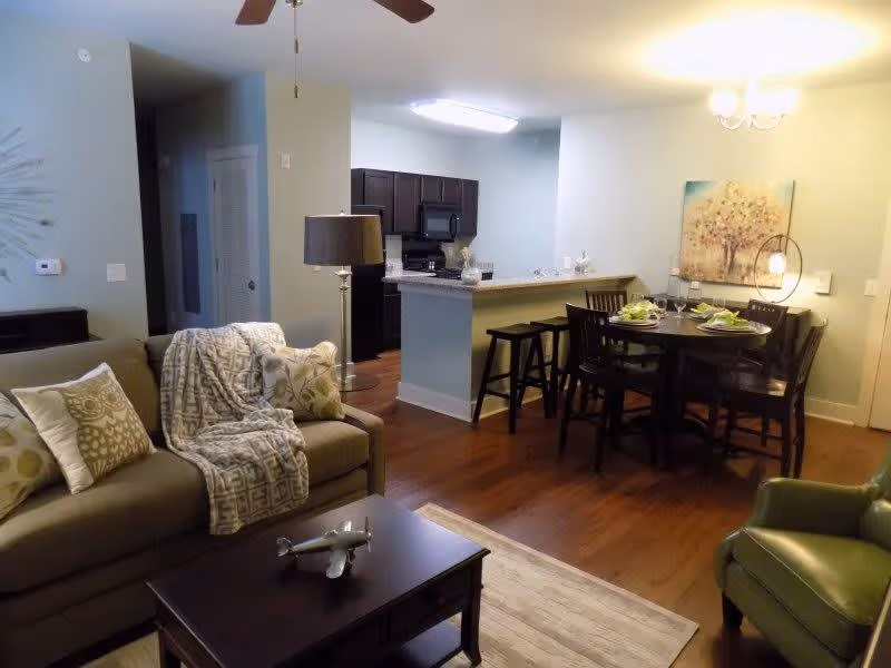 A cozy living and dining area with a beige sofa adorned with patterned pillows and a throw blanket, a dark wooden coffee table with a small airplane model, a green armchair, a dining table set for four with green napkins, and a kitchen area with dark cabinets and a breakfast bar with stools.