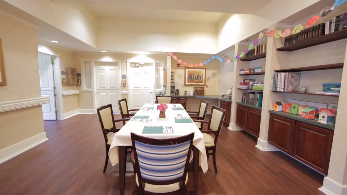 A dining room in an assisted living facility with a rectangular table covered with a white tablecloth, set with placemats, utensils, and a small flower centerpiece. Six chairs surround the table. The room has wooden flooring, beige walls, built-in shelves with books and colorful craft items, and a decorative paper garland hanging across the room.